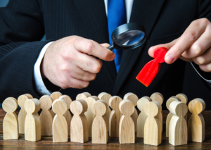 Business person with magnifying glass picking red wooden person from a group of standard wooden figures